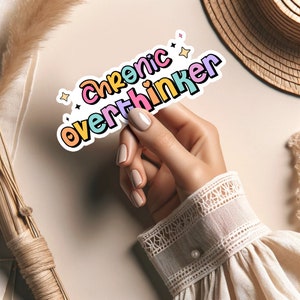 May include: A sticker with the text "Chronic Overthinker" in rainbow colors with stars around it. A hand is holding the sticker.