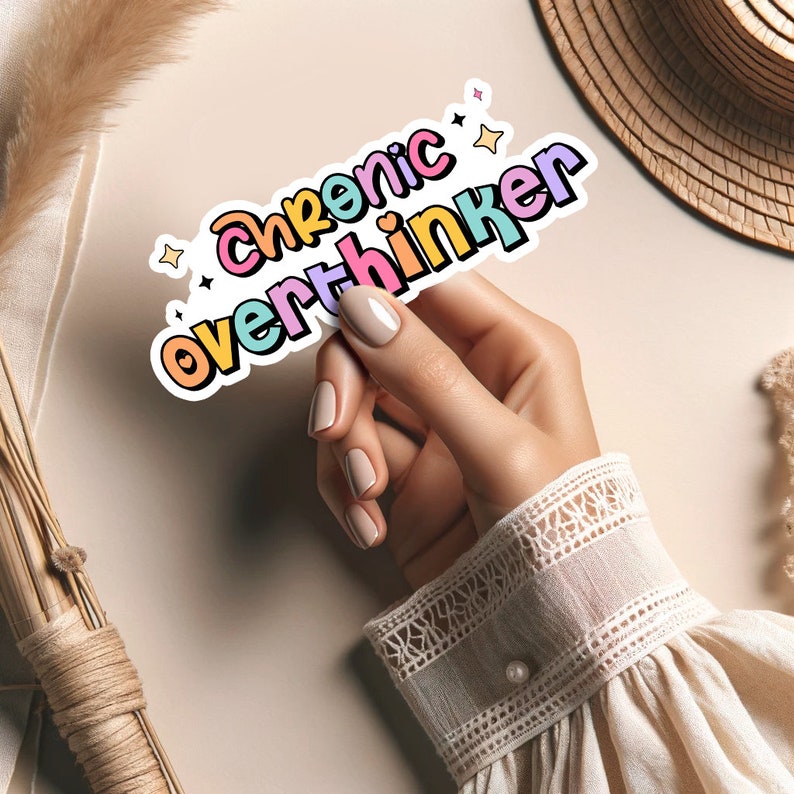May include: A sticker with the text "Chronic Overthinker" in rainbow colors with stars around it. A hand is holding the sticker.