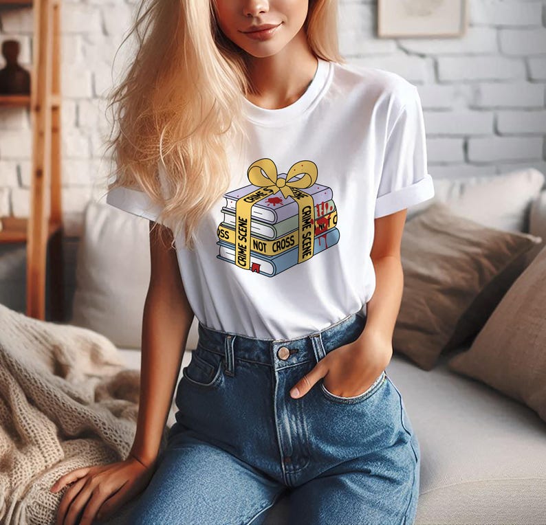 May include: A white t-shirt with a graphic of a stack of books wrapped in yellow crime scene tape with the text "Crime Scene Do Not Cross".