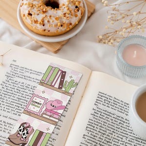 May include: Open book with decorative bookmarks featuring a cowboy theme, a donut, a candle, and a cup of coffee. The bookmarks have illustrations of a cowboy hat, boots, and books. The donut is on a small wooden board.