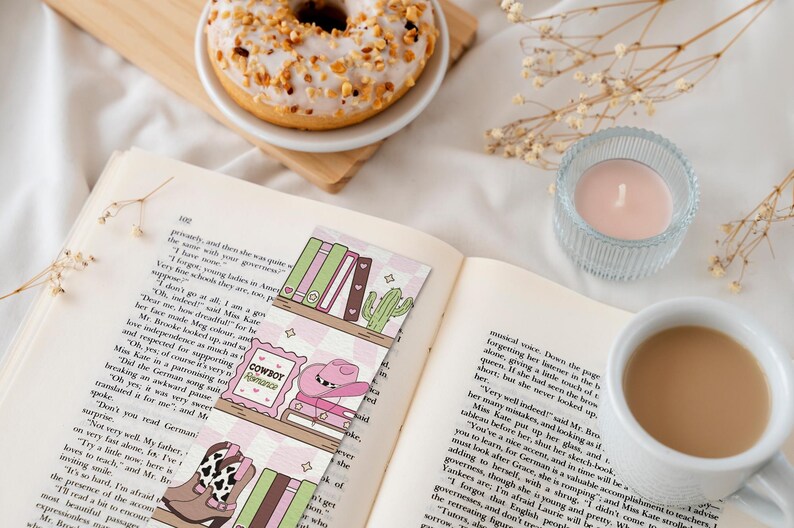 May include: Open book with decorative bookmarks featuring a cowboy theme, a donut, a candle, and a cup of coffee. The bookmarks have illustrations of a cowboy hat, boots, and books. The donut is on a small wooden board.
