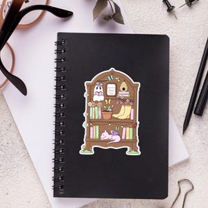 Puede incluir: Una pegatina caprichosa con una estanter&iacute;a marr&oacute;n llena de libros, un fantasma, un drag&oacute;n durmiente y una colmena. El texto "Reading Book Club" es visible. La pegatina est&aacute; sobre un cuaderno espiral negro.