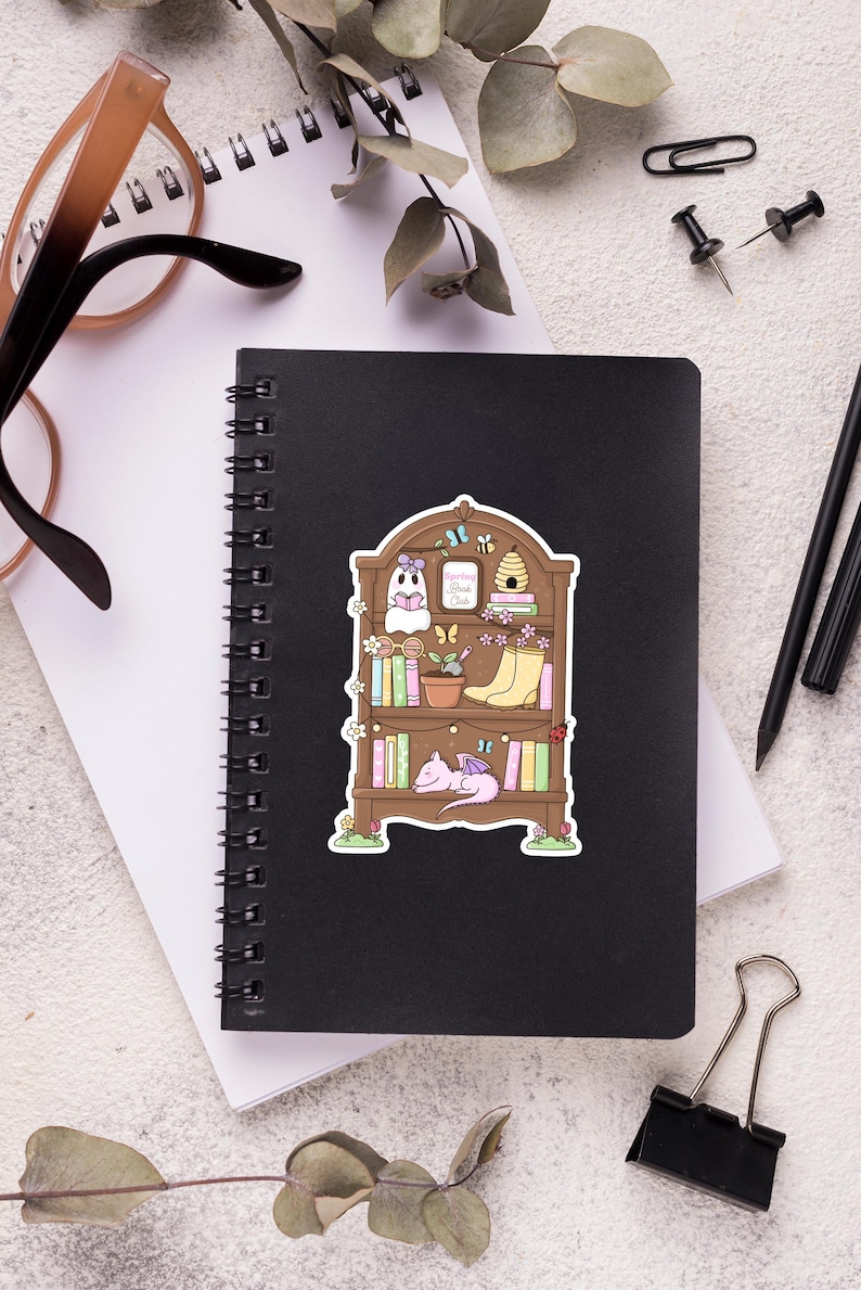 Puede incluir: Una pegatina caprichosa con una estanter&iacute;a marr&oacute;n llena de libros, un fantasma, un drag&oacute;n durmiente y una colmena. El texto "Reading Book Club" es visible. La pegatina est&aacute; sobre un cuaderno espiral negro.