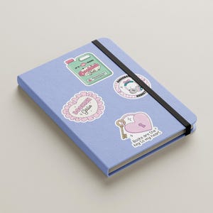 May include: A blue notebook with various stickers on it. The stickers are pink and white with illustrations and text. One sticker says "Bookish Gal", another says "Books are the key to my heart", and another says "Listening Addict".