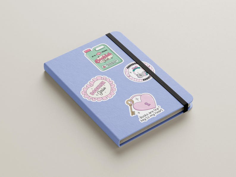 May include: A blue notebook with various stickers on it. The stickers are pink and white with illustrations and text. One sticker says "Bookish Gal", another says "Books are the key to my heart", and another says "Listening Addict".