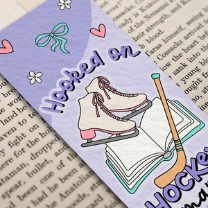 May include: A purple bookmark with a white book, ice skates, and a hockey stick. The bookmark has the text "Hooked on Hockey Romance" in purple letters.