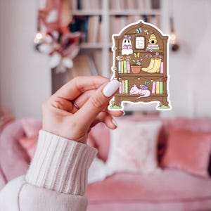Puede incluir: Una pegatina con un dise&ntilde;o de estanter&iacute;a caprichoso. La estanter&iacute;a marr&oacute;n est&aacute; llena de libros coloridos, una planta en maceta y un drag&oacute;n rosa durmiente. La pegatina tambi&eacute;n incluye un fantasma leyendo un libro y el texto "Spring Book Club".
