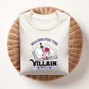 May include: White t-shirt with a graphic design of an open book with a rose and a dagger. The text reads "Falling for the Villain One Page at a Time."