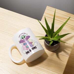May include: White ceramic mug with a cartoon alien in a spaceship holding books. The mug says "Just here for the books".