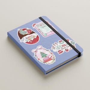 May include: A light blue notebook with a black elastic band, adorned with colorful stickers. Stickers include a Christmas theme, a book-themed design, and the text "Bookman Surprise".