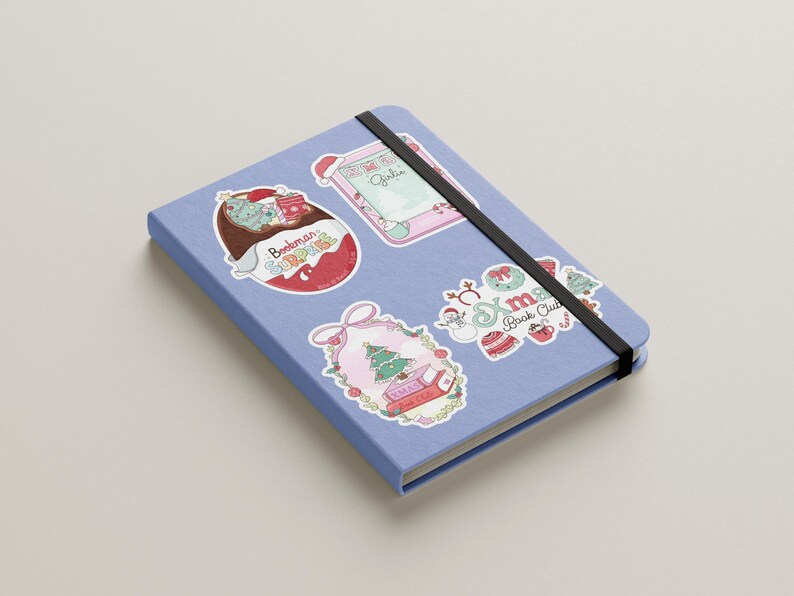 May include: A light blue notebook with a black elastic band, adorned with colorful stickers. Stickers include a Christmas theme, a book-themed design, and the text "Bookman Surprise".