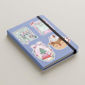 May include: A light blue notebook with a black elastic band closure. The notebook is decorated with several Christmas-themed stickers, including a Christmas tree, a reindeer, and a gingerbread house.