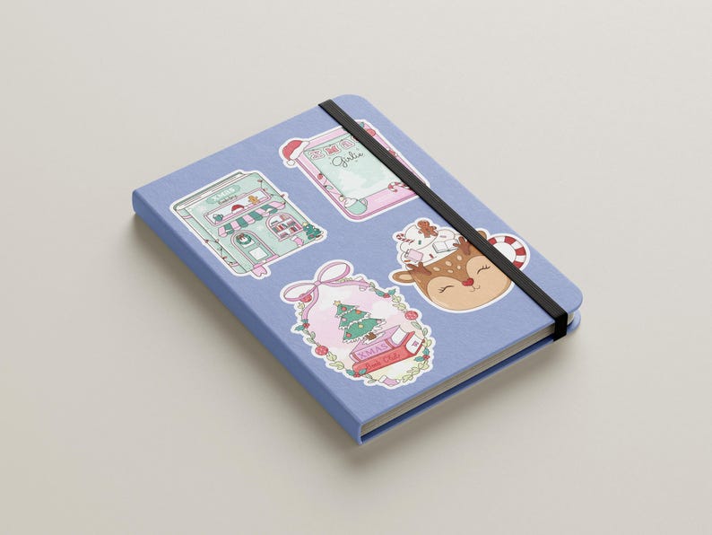 May include: A light blue notebook with a black elastic band closure. The notebook is decorated with several Christmas-themed stickers, including a Christmas tree, a reindeer, and a gingerbread house.