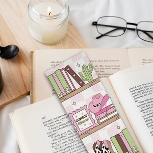 May include: A bookmark with a pink and white checkered pattern featuring a shelf design with books, a cactus, and a cowboy hat. The text "Cowboy Romance" is visible. A black mug of coffee, a candle, and glasses are also in the image.