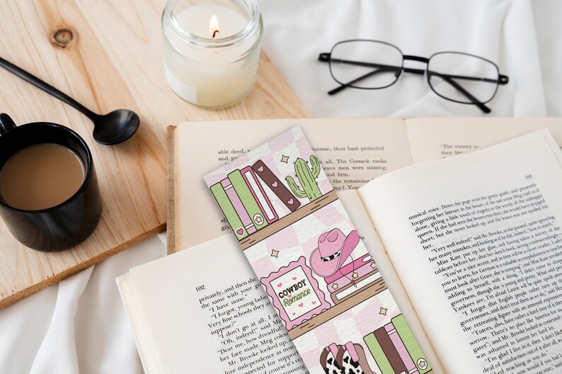 May include: A bookmark with a pink and white checkered pattern featuring a shelf design with books, a cactus, and a cowboy hat. The text "Cowboy Romance" is visible. A black mug of coffee, a candle, and glasses are also in the image.