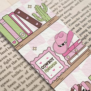 May include: A decorative bookmark featuring a pink cowboy hat, books, and a cactus on a shelf. The bookmark has the text "COWBOY Romance" in a pink frame. The background is a pink and white checkered pattern.