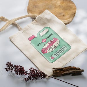 May include: A white canvas tote bag with a green and pink illustration of a bottle labeled "Bookish Fuel" and the text "Up to 24 hours reading. Warning: May Cause Book Hangovers."
