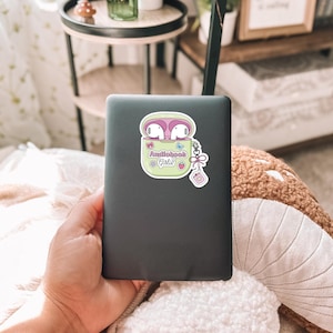 May include: A sticker featuring a cartoon image of white earbuds in a light green case with the text "Audiobook @ Girlie". The sticker has a pink border and is on a dark gray laptop.