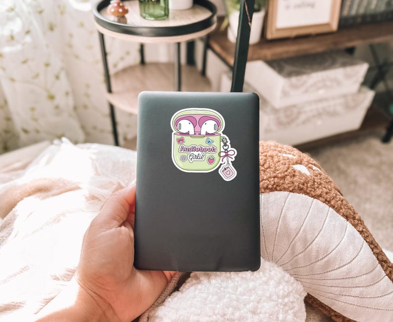 May include: A sticker featuring a cartoon image of white earbuds in a light green case with the text "Audiobook @ Girlie". The sticker has a pink border and is on a dark gray laptop.