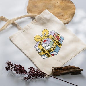 May include: A beige tote bag with a stack of books wrapped in yellow crime scene tape. The books have a red blood stain and the text "Crime Scene Do Not Cross" on the tape.
