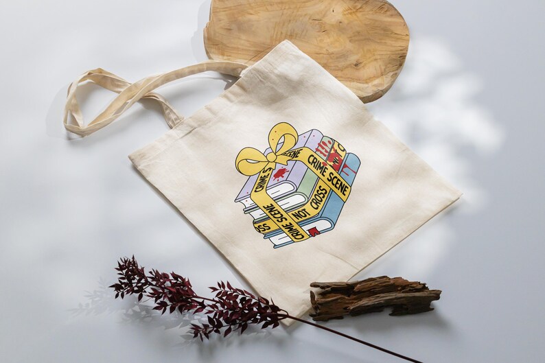 May include: A beige tote bag with a stack of books wrapped in yellow crime scene tape. The books have a red blood stain and the text "Crime Scene Do Not Cross" on the tape.