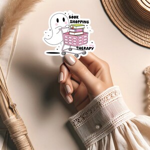 以下が含まれることがあります： 本がいっぱい入ったピンクのショッピングカートを押す白い幽霊。カートには「Book Shopping is my Therapy」と書かれています。