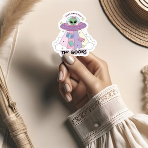 May include: A sticker featuring a cartoon alien in a flying saucer with the text "Just here for the books". The alien is green and has large eyes. The saucer is pink and white with hearts. The books are colorful and are floating around the alien.