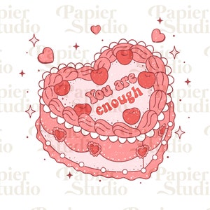 Puede incluir: Un pastel rosa en forma de corazón con glaseado blanco y cerezas rojas. El pastel tiene las palabras "You are enough" escritas en una fuente retro.