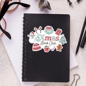 May include: A black spiral-bound notebook with a colorful Christmas-themed sticker that reads "Xmas Book Club." The sticker features a snowman, wreath, sweater, hot cocoa, and gingerbread man. Other items on the white surface include glasses, pens, and a paper clip.
