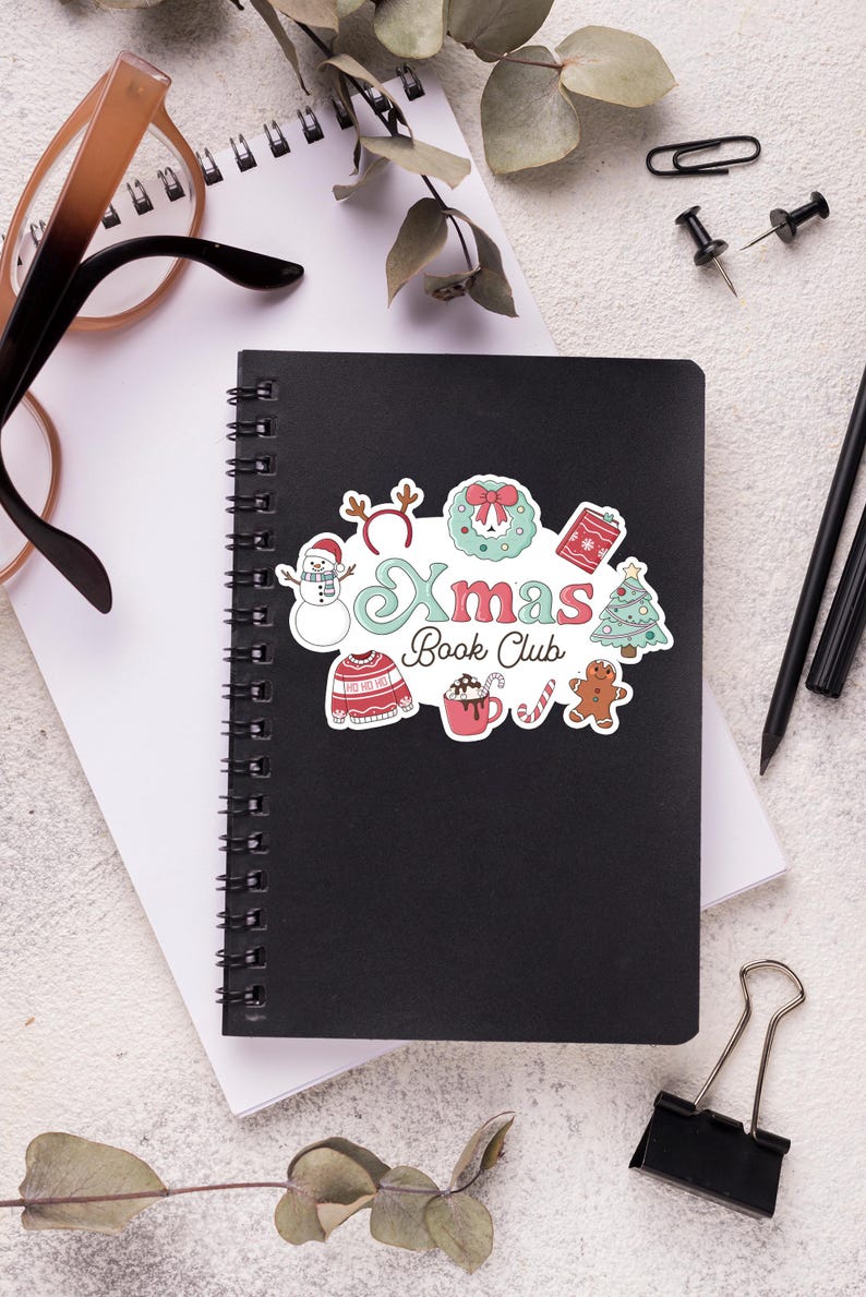 May include: A black spiral-bound notebook with a colorful Christmas-themed sticker that reads "Xmas Book Club." The sticker features a snowman, wreath, sweater, hot cocoa, and gingerbread man. Other items on the white surface include glasses, pens, and a paper clip.