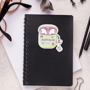 May include: A black spiral-bound notebook with a sticker of a green case containing pink earbuds. The sticker reads "Audiobook Girlie" and features a strawberry and butterfly. The notebook is on a white surface with glasses, pens, and paperclips.