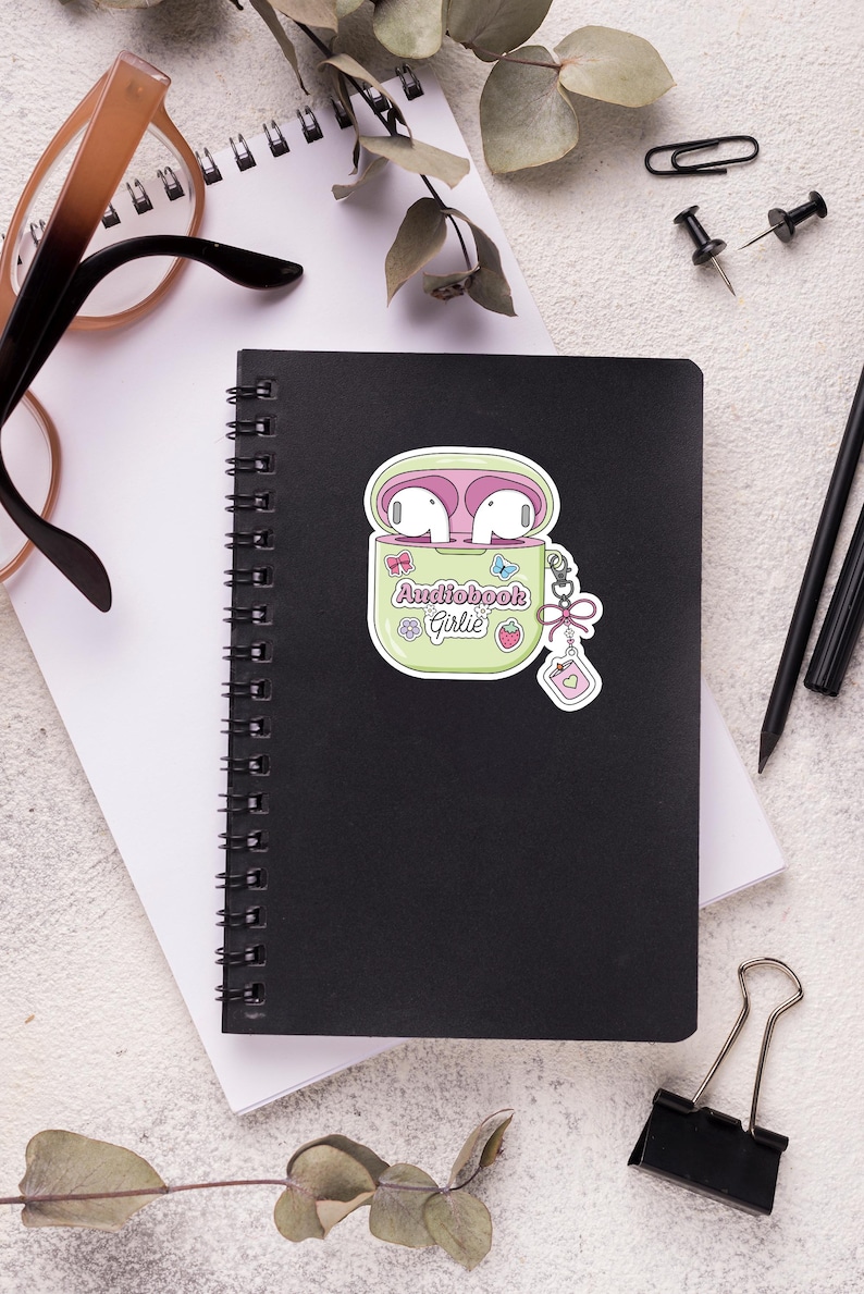 May include: A black spiral-bound notebook with a sticker of a green case containing pink earbuds. The sticker reads "Audiobook Girlie" and features a strawberry and butterfly. The notebook is on a white surface with glasses, pens, and paperclips.