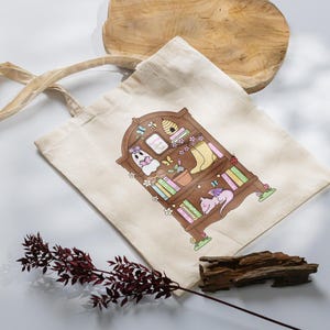 Puede incluir: Bolso tote beige con una ilustraci&oacute;n caprichosa de una estanter&iacute;a de madera llena de libros, un fantasma, un drag&oacute;n durmiente y otros elementos decorativos. El bolso tiene asas largas y est&aacute; hecho de una tela resistente.