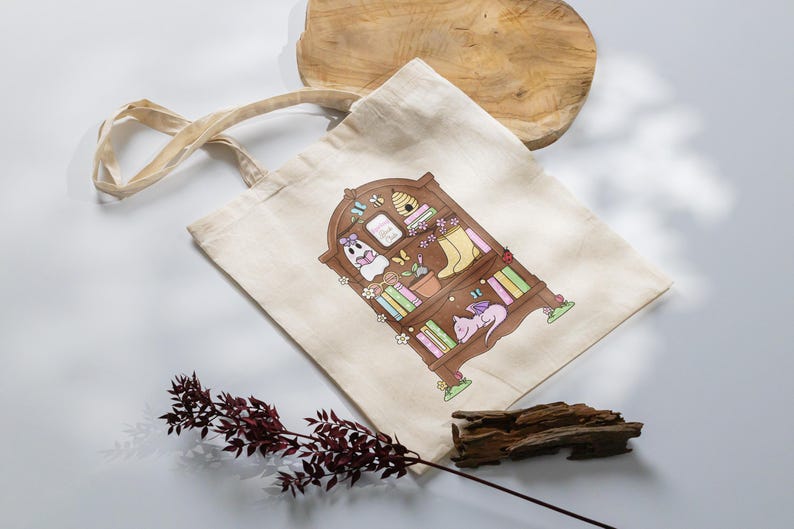 Puede incluir: Bolso tote beige con una ilustraci&oacute;n caprichosa de una estanter&iacute;a de madera llena de libros, un fantasma, un drag&oacute;n durmiente y otros elementos decorativos. El bolso tiene asas largas y est&aacute; hecho de una tela resistente.