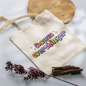 May include: A beige tote bag with a colorful graphic that says "chronic overthinker".