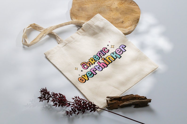 May include: A beige tote bag with a colorful graphic that says "chronic overthinker".