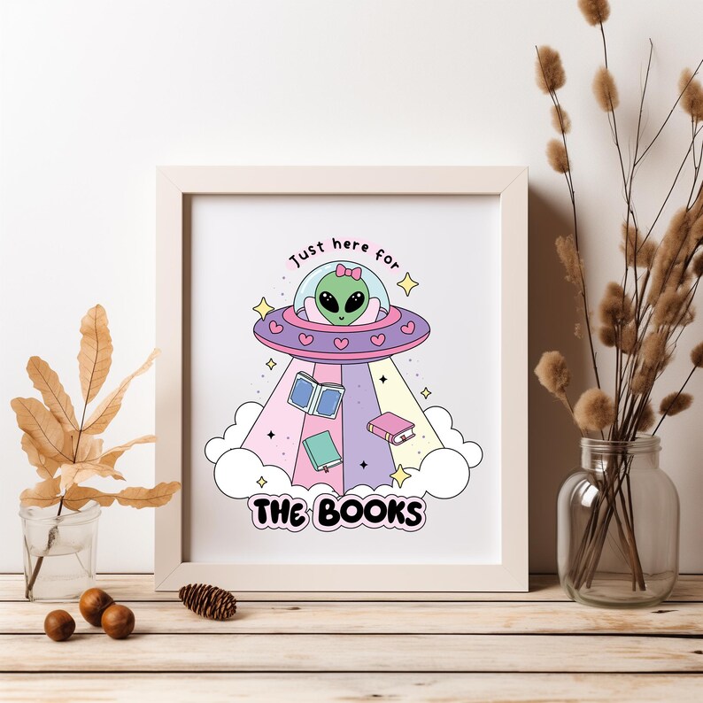 May include: A white framed print with a cartoon illustration of a green alien in a pink and purple spaceship with the text "Just here for the books" and stars.