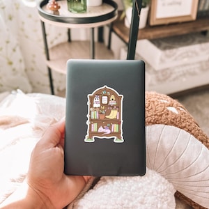 Puede incluir: Una tableta negra con una pegatina colorida de una estanter&iacute;a. La pegatina presenta libros ilustrados, plantas, un fantasma, una colmena y un gato durmiendo. La tableta est&aacute; sujeta en la mano de una persona.