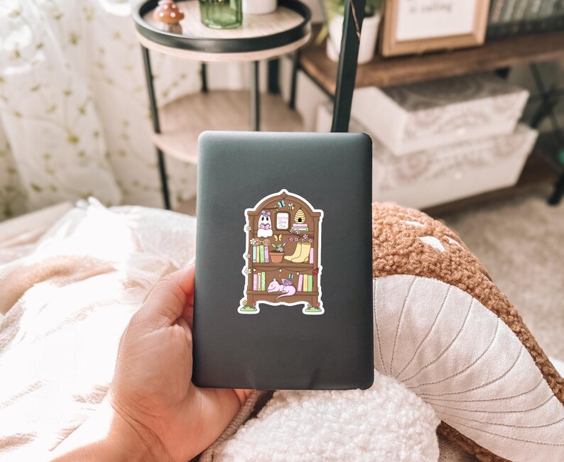 Puede incluir: Una tableta negra con una pegatina colorida de una estanter&iacute;a. La pegatina presenta libros ilustrados, plantas, un fantasma, una colmena y un gato durmiendo. La tableta est&aacute; sujeta en la mano de una persona.