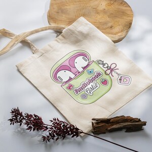May include: A beige tote bag with a graphic of a green and pink AirPod case, with the text "Audiobook Girlie". The bag has a matching keychain with a strawberry and heart charm. The bag is on a white surface.