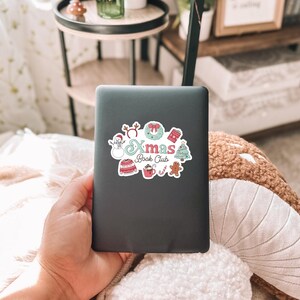 May include: A black e-reader with a colorful sticker that says "Xmas Book Club." The sticker features festive illustrations of a snowman, wreath, sweater, hot cocoa, gingerbread man, and Christmas tree. The e-reader is held in a person's hand.
