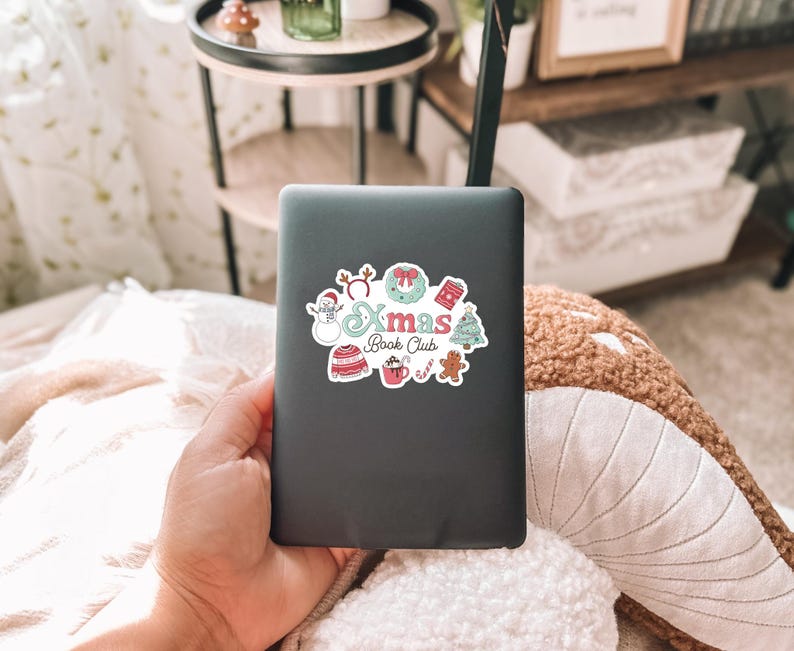 May include: A black e-reader with a colorful sticker that says "Xmas Book Club." The sticker features festive illustrations of a snowman, wreath, sweater, hot cocoa, gingerbread man, and Christmas tree. The e-reader is held in a person's hand.