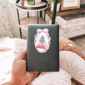 May include: A sticker featuring a Christmas tree atop two books with the text "XMAS Book Club" inside a pink and green floral frame, topped with a pink bow. The sticker is on a dark gray laptop.