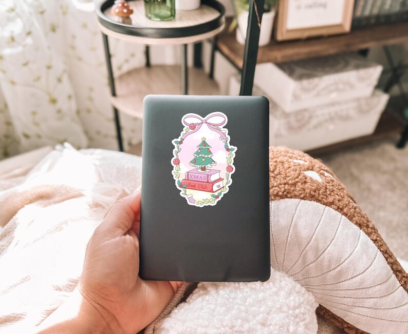 May include: A sticker featuring a Christmas tree atop two books with the text "XMAS Book Club" inside a pink and green floral frame, topped with a pink bow. The sticker is on a dark gray laptop.