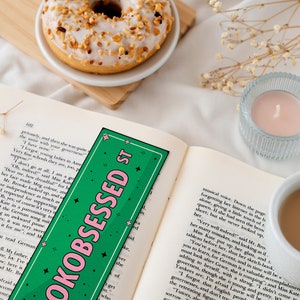 May include: A green and pink bookmark with the text "OKOBSESSED ST" in pink letters. The bookmark is on top of an open book with a cup of coffee and a donut in the background.