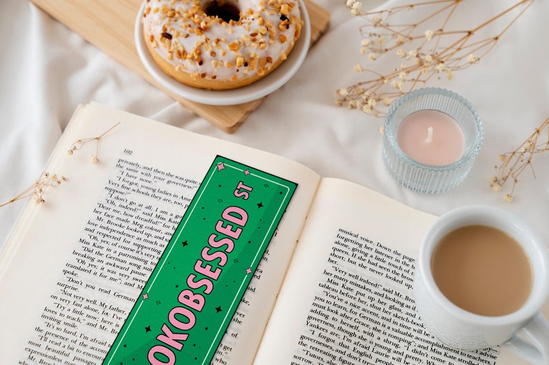 May include: A green and pink bookmark with the text "OKOBSESSED ST" in pink letters. The bookmark is on top of an open book with a cup of coffee and a donut in the background.