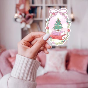 May include: A sticker featuring a Christmas tree atop two books with the text "XMAS Book Club." The design is framed by a pink bow and a wreath of leaves and berries, with a pink background. A small stocking is also included in the design.