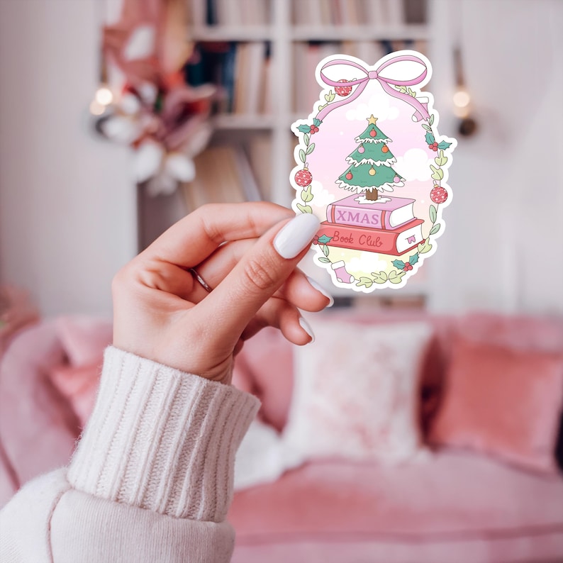 May include: A sticker featuring a Christmas tree atop two books with the text "XMAS Book Club." The design is framed by a pink bow and a wreath of leaves and berries, with a pink background. A small stocking is also included in the design.