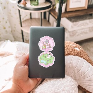 Pu&ograve; includere: Un laptop nero con due adesivi rosa a forma di nuvola. L'adesivo superiore raffigura una stanza con una sedia e dei libri. L'adesivo inferiore mostra un personaggio dei cartoni animati in un parco. Il laptop &egrave; tenuto in mano.