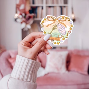 May include: Heart-shaped sticker with a key, books, and the text "Key to Stars Hollow Library" and "Autumn Book Club." The sticker has a pastel color scheme with a bow and floral accents. The sticker is held by a person.